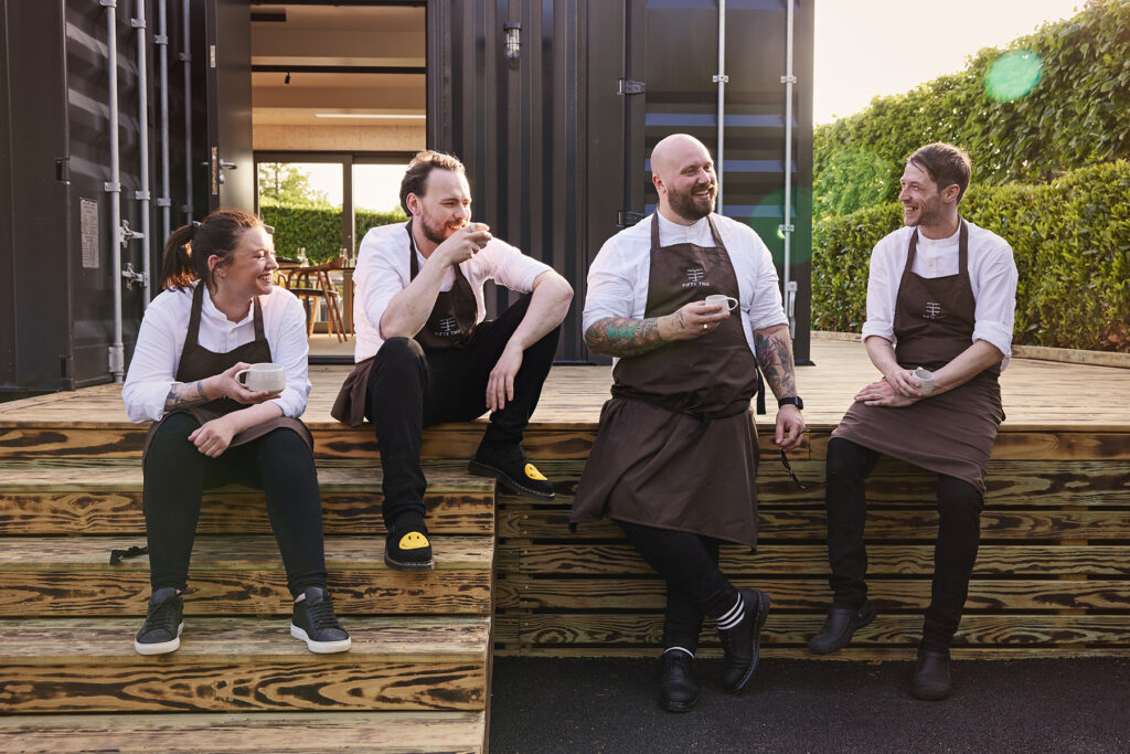 adam-degg-head-chef-and-team-enjoy-coffee-outside restaurant steps timber deck steel container doors