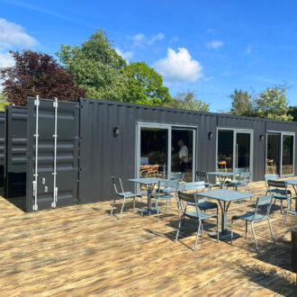 external view timber deck grey shipping container al fresco dining area planters grey tables chairs blue sky trees
