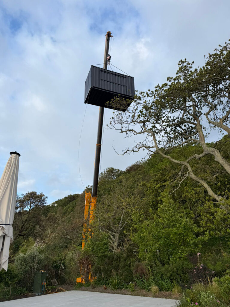 Large yellow crane lifting 20ft converted shipping container catering unit over trees onto concrete slab